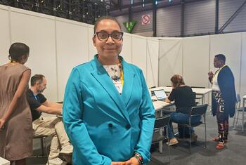 Una mujer con chaqueta azul y gafas se encuentra de pie en un evento de una conferencia, con otros asistentes al fondo.