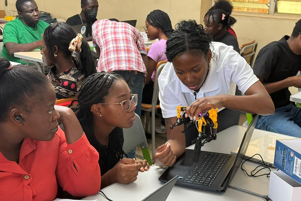 A group of students in a classroom setting collaborates around a laptop, with one student assembling a small yellow and black robotic arm.