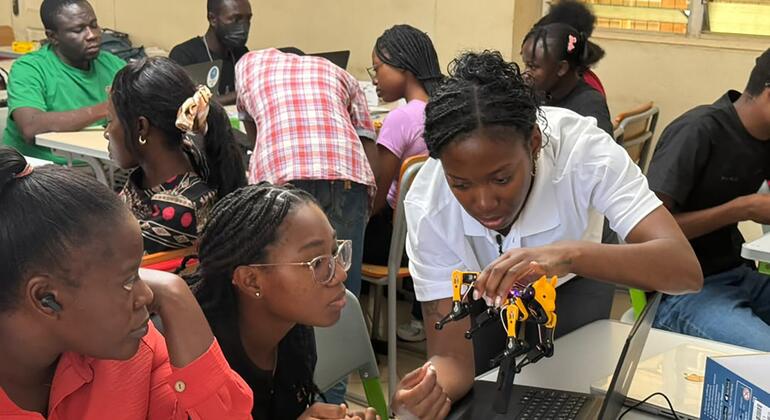 A group of students in a classroom setting collaborates around a laptop, with one student assembling a small yellow and black robotic arm.