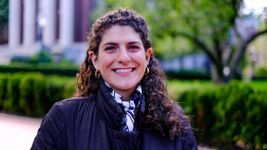 A portrait of a woman with curly brown hair smiling at the camera. She is wearing a black jacket and a black and white patterned scarf, with gold hoop earrings. The background is an out-of-focus outdoor scene with green bushes and a building.