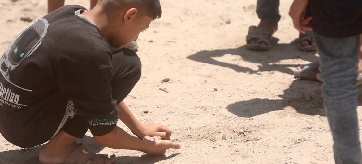 Un niño pequeño está en cuclillas en la arena jugando con canicas de cristal en Gaza.
