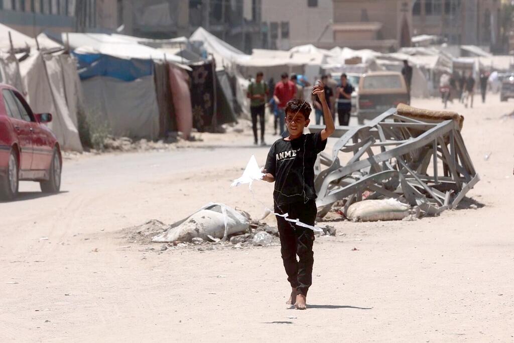 Un enfant palestinien marche pieds nus, portant un cerf-volant artisanal, à travers les décombres d'Al-Rimal, dans la ville de Gaza, avec un campement de tentes en arrière-plan.