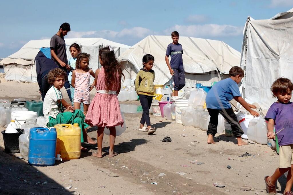 Dans un camp de réfugiés, des enfants et des jeunes font la queue avec différents récipients pour recevoir de l'eau d'un camion de livraison.