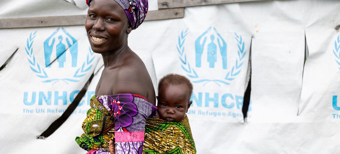 Mwanamke wa Sudan Kusini mwenye tabasamu amebeba mtoto mgongoni, amesimama mbele ya hema la UNHCR katika kambi ya wakimbizi.