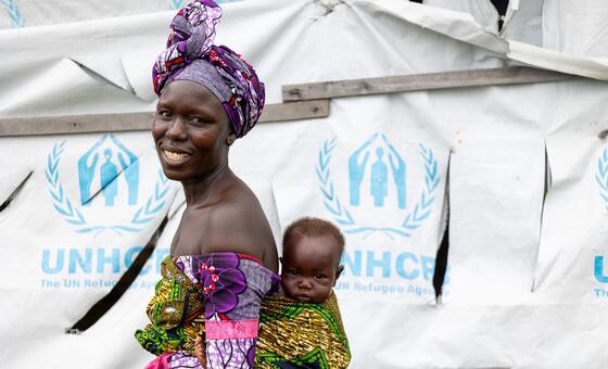 Mwanamke wa Sudan Kusini mwenye tabasamu amebeba mtoto mgongoni, amesimama mbele ya hema la UNHCR katika kambi ya wakimbizi.