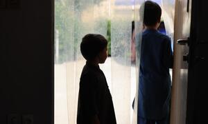 دو نوجوان لڑکے، ایک سیاہ قمیض میں اور دوسرا نیلے رنگ کی شلوار قمیض میں، پردوں سے کھڑکی سے باہر دیکھ رہے ہیں۔