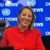 Uma mulher de jaqueta vermelha sorri enquanto está sentada em frente a um microfone em um estúdio com o logotipo da ONU News ao fundo.