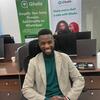 Mwanamume mwenye tabasamu akiwa amevaa suti ya biashara ameketi kwenye dawati ofisini, huku mabango ya chapa ya Ghala na chupa ya maji yakionekana nyuma.