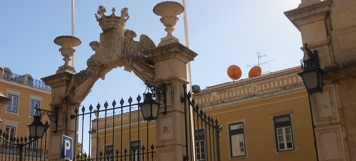 Portão de pedra ornamentado do edifício PALCO em Portugal, com um prédio amarelo ao fundo sob um céu azul claro.