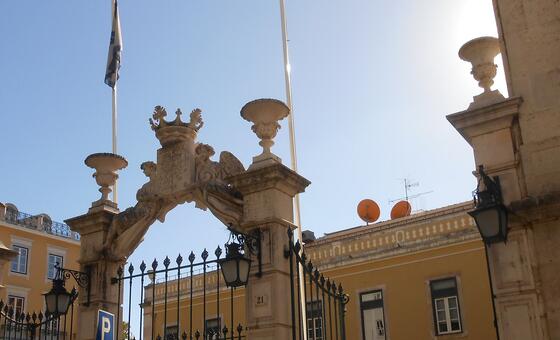 Portão de pedra ornamentado do edifício PALCO em Portugal, com um prédio amarelo ao fundo sob um céu azul claro.