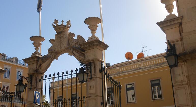 Portão de pedra ornamentado do edifício PALCO em Portugal, com um prédio amarelo ao fundo sob um céu azul claro.