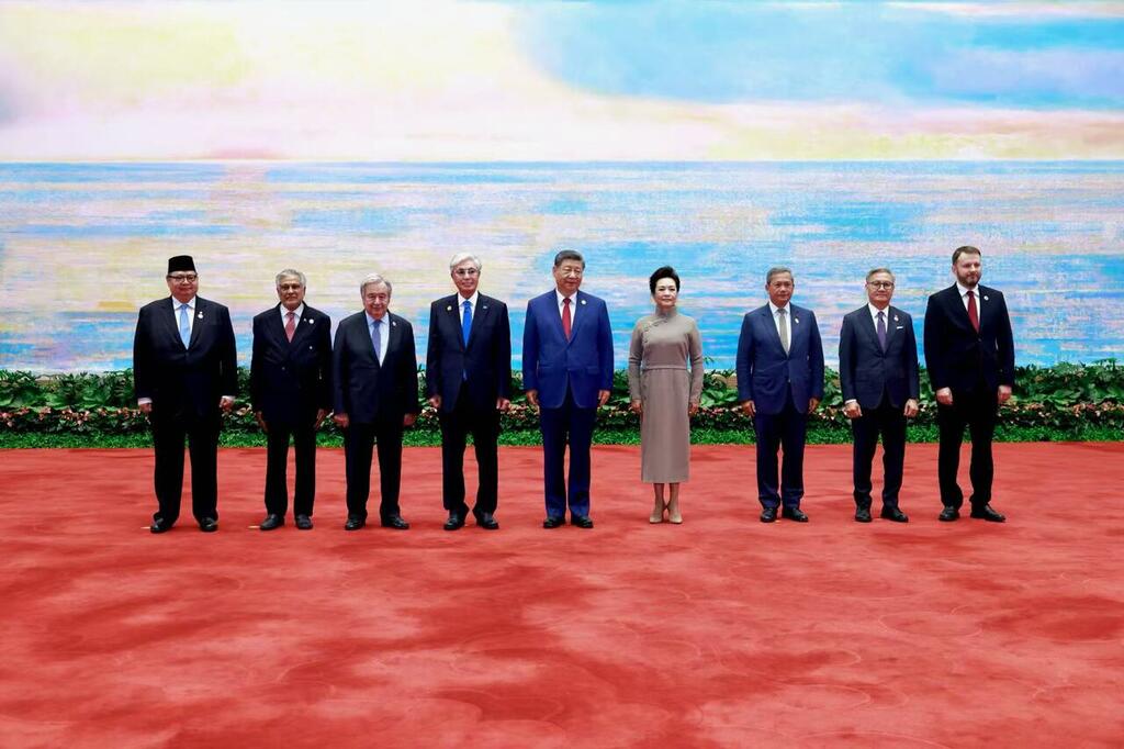 Un groupe de dirigeants mondiaux et leurs conjoints posent pour une photo sur un tapis rouge devant un grand écran.