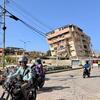 Motorcyclists ride down a street in La Guaira, Venezuela, with damaged buildings and a collapsed structure visible in the background.