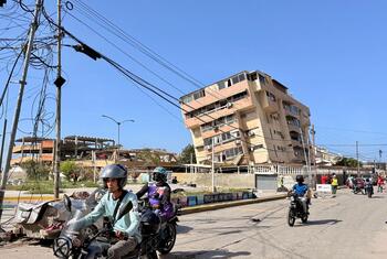 Motociclistas circulan por una calle de La Guaira, Venezuela, con edificios dañados y una estructura derrumbada visibles al fondo.