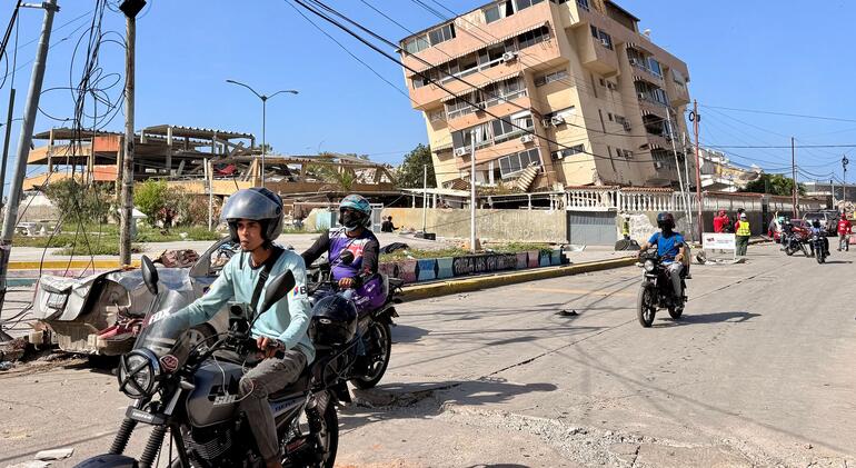Motociclistas circulan por una calle de La Guaira, Venezuela, con edificios dañados y una estructura derrumbada visibles al fondo.