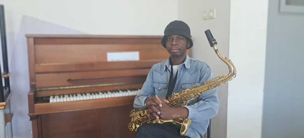Um homem negro, vestindo camisa jeans e chapéu, segura um saxofone enquanto está sentado em frente a um piano.