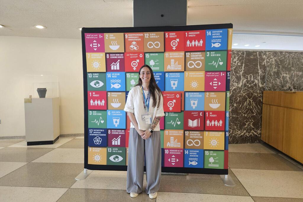 Una mujer sonriente, vestida con una blusa blanca y pantalones grises, posa frente a una colorida exposición sobre los Objetivos de Desarrollo Sostenible de las Naciones Unidas dentro de un edificio.