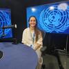Una mujer de pelo largo y oscuro y camisa blanca está sentada en una mesa azul con un micrófono en un estudio, con logotipos de la ONU en pantallas detrás de ella.