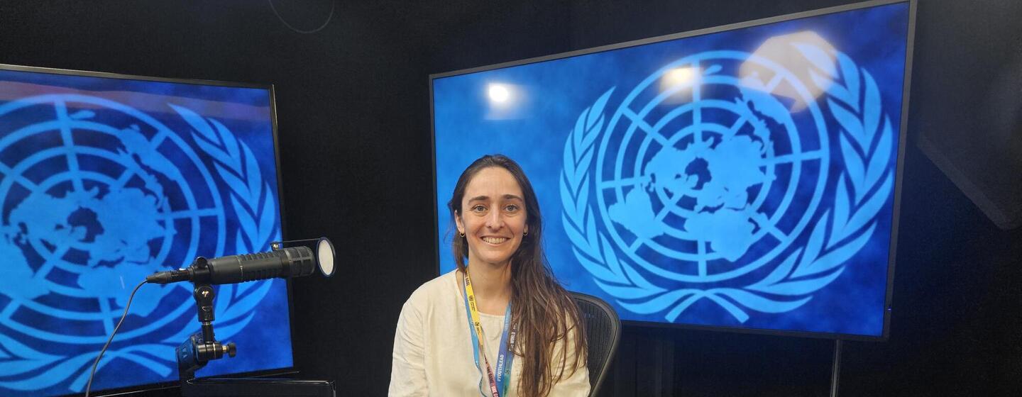 Una mujer de pelo largo y oscuro y camisa blanca está sentada en una mesa azul con un micrófono en un estudio, con logotipos de la ONU en pantallas detrás de ella.