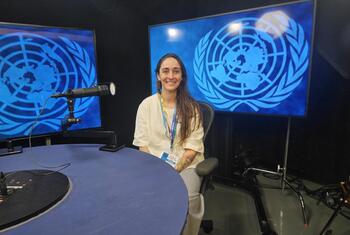 Una mujer de pelo largo y oscuro y camisa blanca está sentada en una mesa azul con un micrófono en un estudio, con logotipos de la ONU en pantallas detrás de ella.