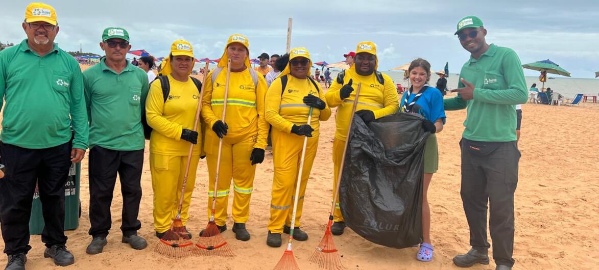 Um grupo de voluntários com uniformes amarelos e verdes posa em uma praia de areia com rastelos e um grande saco de lixo para um evento de limpeza.