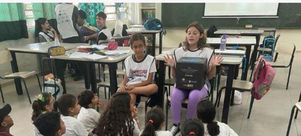 Uma menina está em pé numa sala de aula, segurando uma caixa preta e falando com um grupo de crianças sentadas.