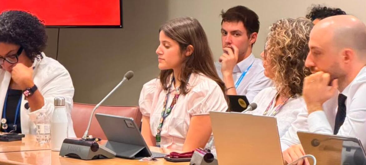 Um grupo de cinco pessoas, das quais quatro são visíveis, está sentado em uma longa mesa de madeira para conferências, com laptops e microfones, durante uma mesa redonda ou reunião.