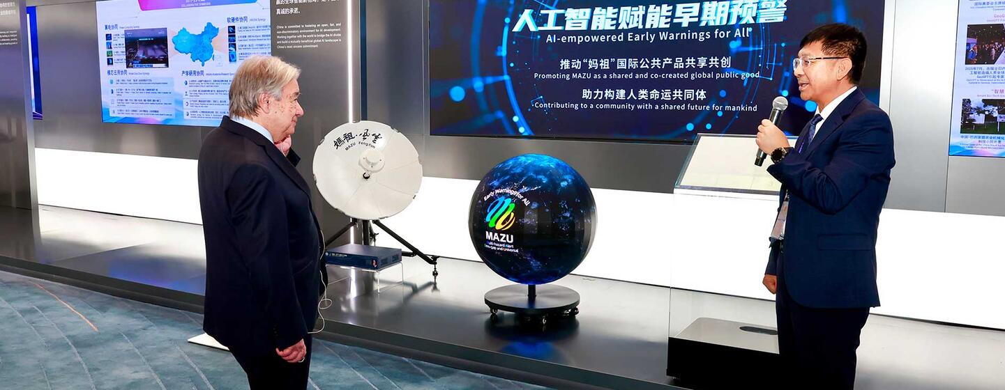 在一个科技展览会上，一名身穿西装的男子对着麦克风讲话，另一名男子在一旁聆听，旁边的大屏幕上显示着人工智能驱动的气象预警系统。