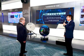 En una exposición de tecnología, un hombre de traje habla por un micrófono mientras otro escucha; una gran pantalla muestra un sistema de alerta temprana meteorológica basado en inteligencia artificial.