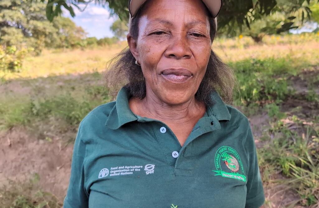 Mwanamke aliyevaa shati la polo la kijani lenye nembo za Shirika la Chakula na Kilimo la UMOJA WA MATAIFA na Huduma ya Misitu Tanzania, akiwa amesimama nje.
