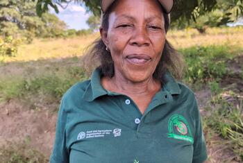 Mwanamke aliyevaa shati la polo la kijani lenye nembo za Shirika la Chakula na Kilimo la UMOJA WA MATAIFA na Huduma ya Misitu Tanzania, akiwa amesimama nje.