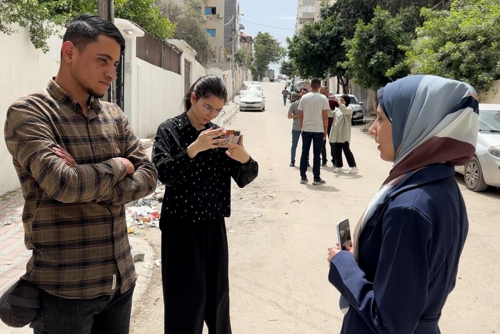 امرأة ترتدي الحجاب تحمل هاتفًا ذكيًا أثناء حديثها مع شخصين آخرين في أحد شوارع غزة خلال جلسة تدريبية على الصحافة المتنقلة.