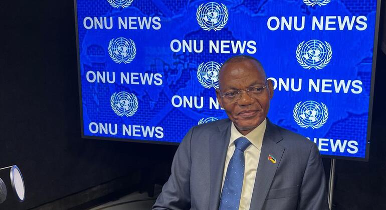 Um homem de terno e gravata está sentado em um estúdio em frente a uma tela azul com a inscrição 'ONU NEWS'.