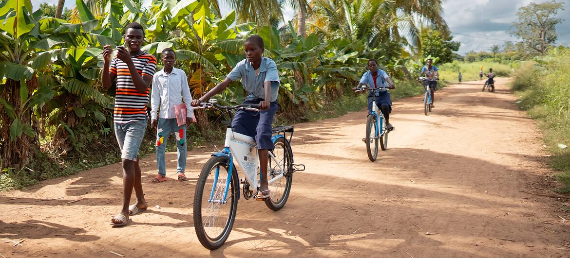 Crianças uniformizadas andam de bicicleta em uma estrada de terra em uma área rural de Moçambique, com palmeiras e vegetação exuberante ao fundo.