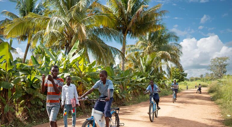 Crianças uniformizadas andam de bicicleta em uma estrada de terra em uma área rural de Moçambique, com palmeiras e vegetação exuberante ao fundo.