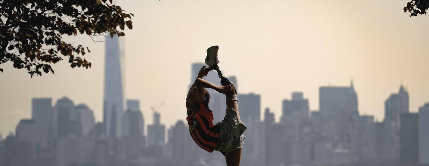 A woman in a striped shirt and shorts performs a high kick pose with a city skyline in the background.