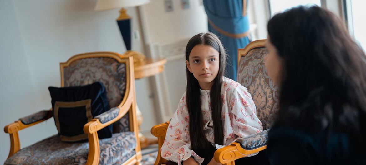 A young Ukrainian girl in a floral blouse sits in an ornate chair and talks to a woman across from her.