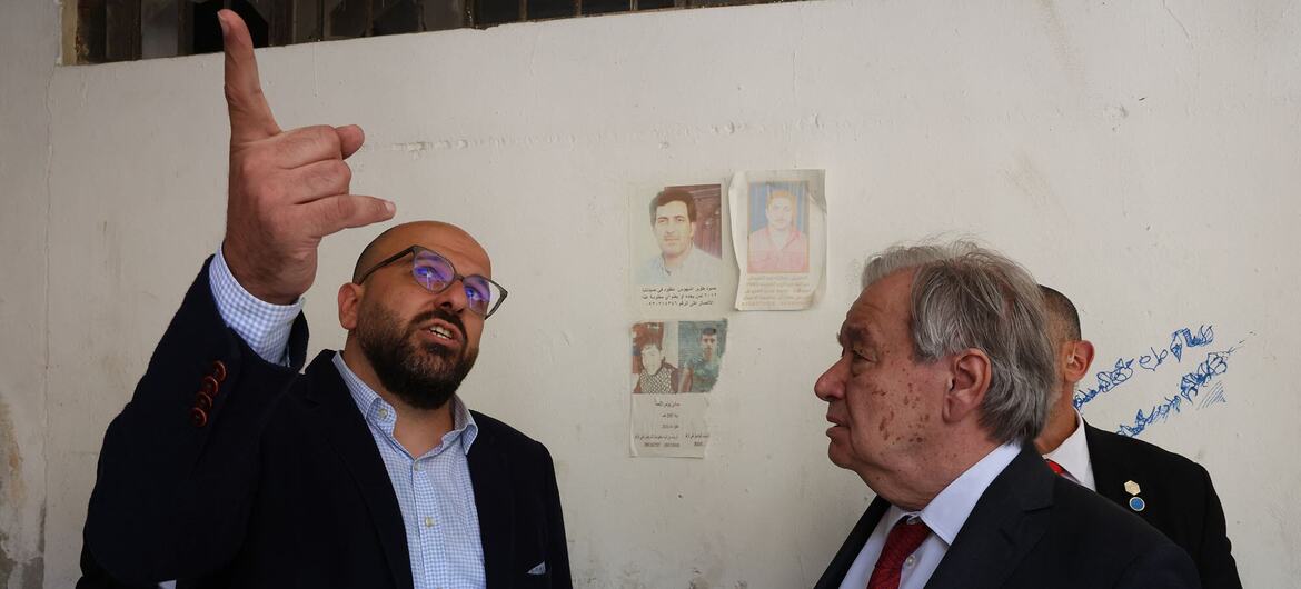 A bald, bearded man in glasses points upwards while talking to an older man with grey hair in a suit and red tie, in front of a white wall with photos and Arabic writing.