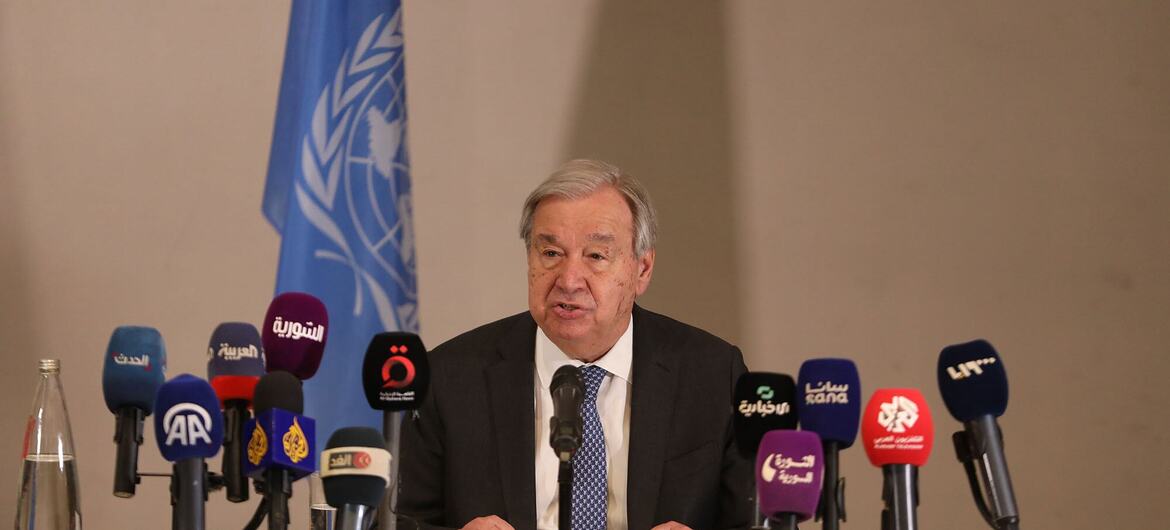 UN Secretary-General speaks at a podium during a press conference in Damascus, with a UN flag in the background and multiple microphones from various news agencies.