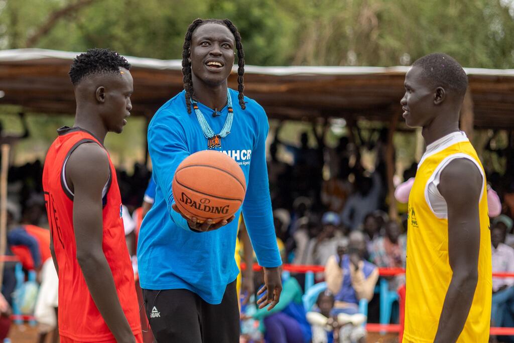 Wenyen Gabriel, ambassadeur de bonne volonté du HCR et joueur de basket-ball professionnel, sourit en tenant un ballon de basket tout en discutant avec deux jeunes hommes sur un terrain de basket dans un camp de réfugiés du Sud-Soudan.