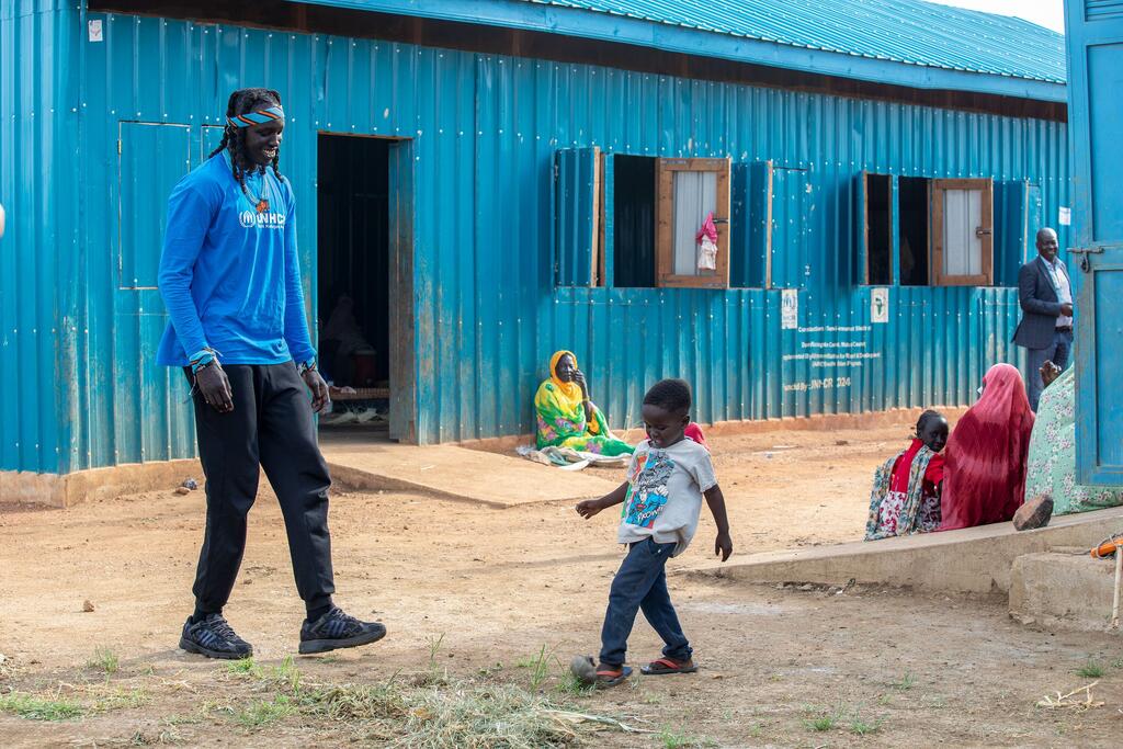 L’ambassadrice de bonne volonté du HCR, Wenyen Gabriel, marche avec un jeune garçon dans un camp de réfugiés au Soudan du Sud. À l’arrière-plan, un bâtiment en tôle ondulée bleue abrite d’autres personnes.