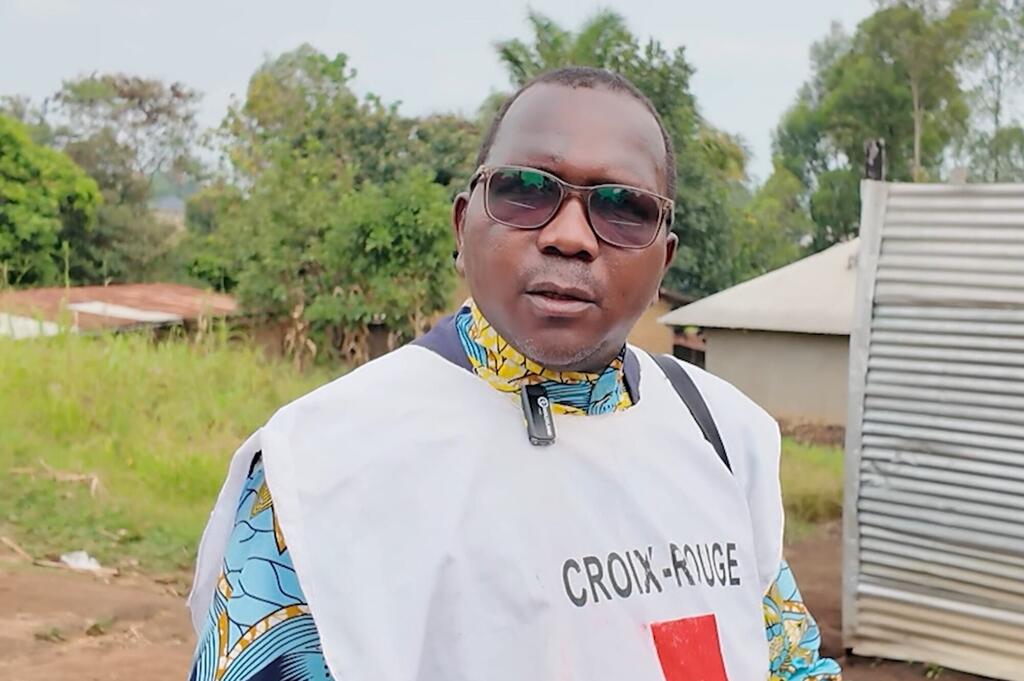 Mwanamume aliyevaa fulana nyeupe yenye nembo ya 'CROIX-ROUGE' amesimama nje katika mazingira ya vijijini, pengine wakati wa kampeni ya uhamasishaji kuhusu Ebola huko Ituri, DRC.