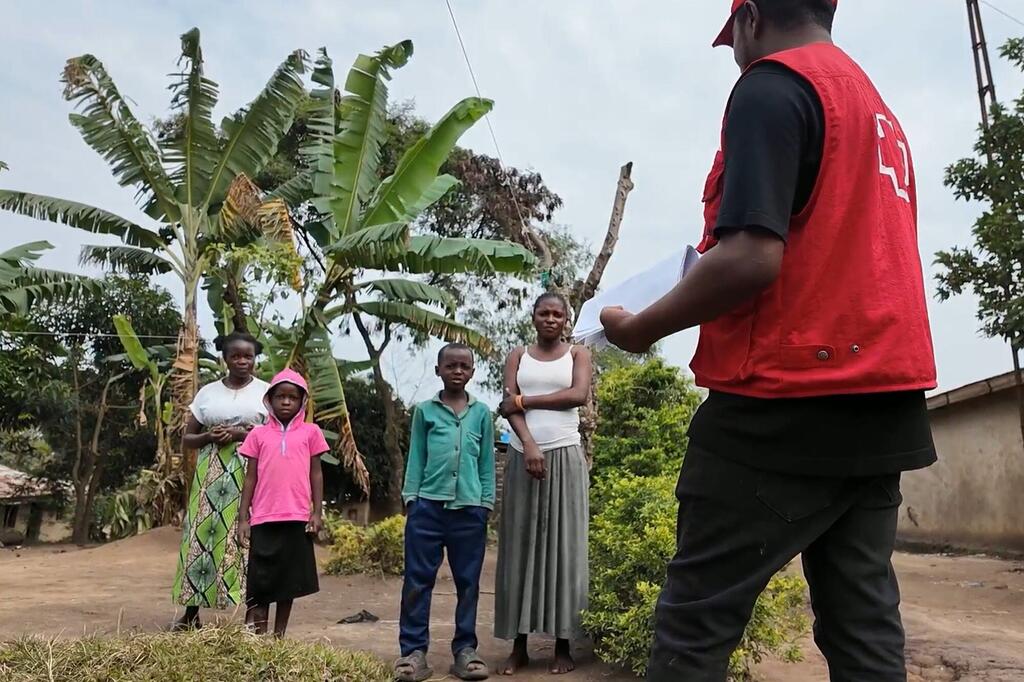 Un travailleur humanitaire vêtu d'un gilet rouge s'adresse à un groupe de quatre personnes, dont deux femmes et deux enfants, dans un cadre rural avec des bananiers en arrière-plan.