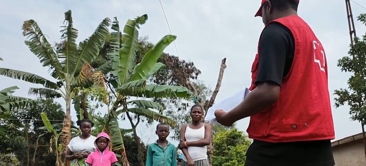 Mfanyakazi wa misaada aliyevaa fulana nyekundu anazungumza na kundi la watu wanne, wakiwemo wanawake wawili na watoto wawili, katika mazingira ya vijijini yenye miti ya ndizi nyuma.