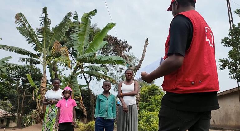 Mfanyakazi wa misaada aliyevaa fulana nyekundu anazungumza na kundi la watu wanne, wakiwemo wanawake wawili na watoto wawili, katika mazingira ya vijijini yenye miti ya ndizi nyuma.