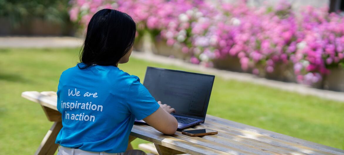 Uma mulher de cabelos escuros e camiseta azul está sentada em uma mesa de piquenique ao ar livre, usando um laptop. A camiseta tem a frase "Somos inspiração em ação" estampada nas costas.