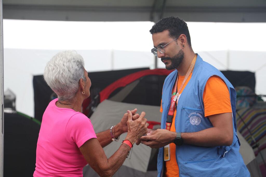 Una anciana con una camisa rosa toma de la mano a un cooperante masculino que lleva un chaleco azul del ACNUR , en lo que parece ser un campamento de ayuda provisional.