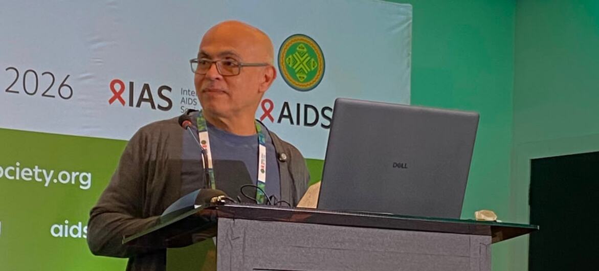 Um homem careca de óculos discursando em um pódio durante uma conferência sobre AIDS em 2026.
