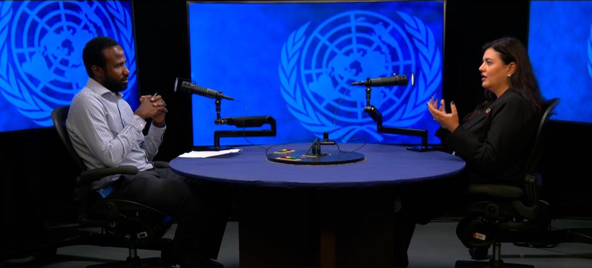 رجل وامرأة يجلسان على طاولة مستديرة مزودة بميكروفونات في استوديو، مع عرض شعارات United Nations على شاشات في الخلفية.