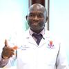 Daktari wa kiume aliyevaa koti jeupe lenye lebo ya jina na nembo ya hospitali anaelekeza kamera.